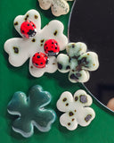 A close-up of several flower-shaped decorative items arranged on a green surface, including white clover with black spots, a green-and-white marbled clover, a teal clover, and two red ladybug embellishments on a white clover. A dark reflective circular object is partially visible on the right.