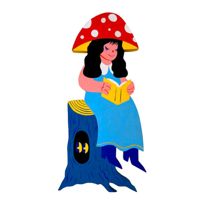 Die cut painted wooden sculpture of a woman in a blue dress and a red toadstool mushroom cap as a hat. She reads a book and sits on a tree trunk, which has a pair of yellow eyes peering off to the side within it.