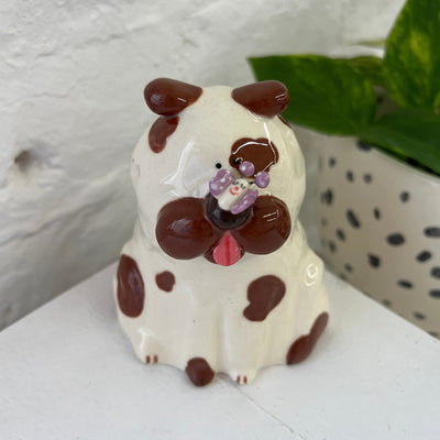 Glazed ceramic sculpture of a white dog with brown spots. It sits with its tongue out and atop its nose is a small, cute purple butterfly with a smiling face.