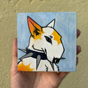 A hand holds a square wood panel with a stylized illustration of a white dog with orange and yellow patches, black eyes, and gray spikes around its neck, against a light blue painted background. 