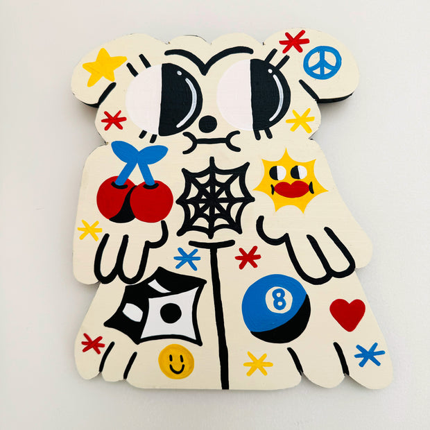 Die cut cream colored wooden painting of a dog with very large cartoony eyes and primary colored tattoos. Tattoos are of cherries, stars, 8 ball and dice.
