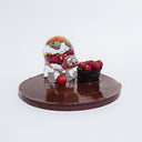 Clear resin sculpture of a quadruped creature with minimal features, and a rotund body. Inside its body are many small fruits with comical faces. It stands next to a basket of apples with similar comical faces. Both are on a brown glitter circular base.