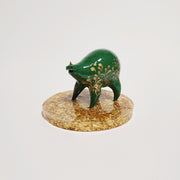 Small sculpture of a green quadruped creature with a small face and very rotund body. It has a single tiny red horn and gold painted patterns along its side. It stands on a gold speckled clear base.