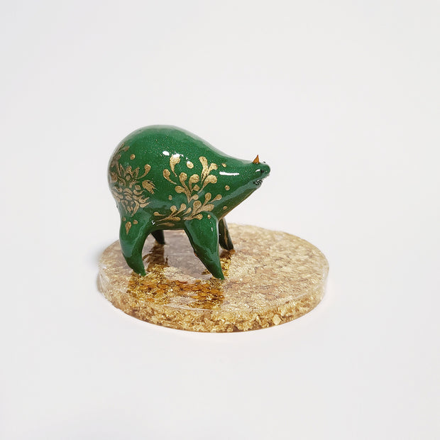Small sculpture of a green quadruped creature with a small face and very rotund body. It has a single tiny gold horn and gold painted patterns along its side. It stands on a gold speckled clear base.