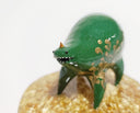 Small sculpture of a green quadruped creature with a small face and very rotund body. It has a single tiny gold horn and gold painted patterns along its side. It stands on a gold speckled clear base.