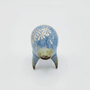Rounded sage blue ceramic sculpture of a quadruped with short legs and small silver eyes over a goofy open mouth. Its body features a silver daisy pattern.