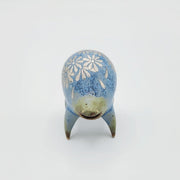 Rounded sage blue ceramic sculpture of a quadruped with short legs and small silver eyes over a goofy open mouth. Its body features a silver daisy pattern.