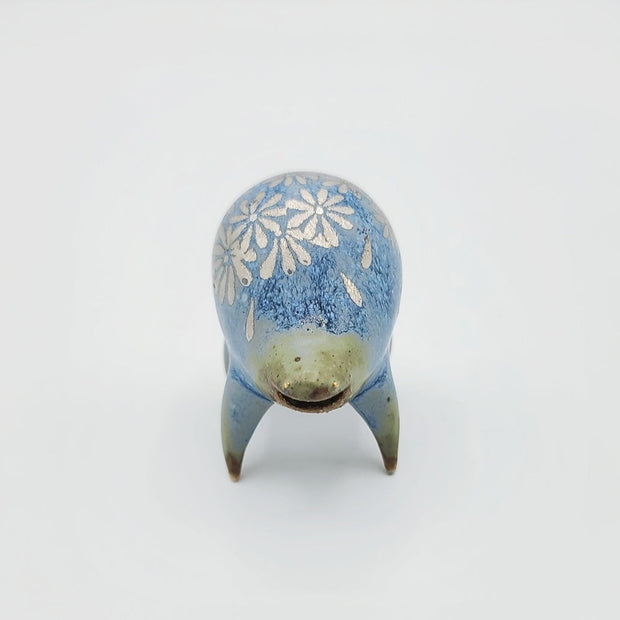 Rounded sage blue ceramic sculpture of a quadruped with short legs and small silver eyes over a goofy open mouth. Its body features a silver daisy pattern.