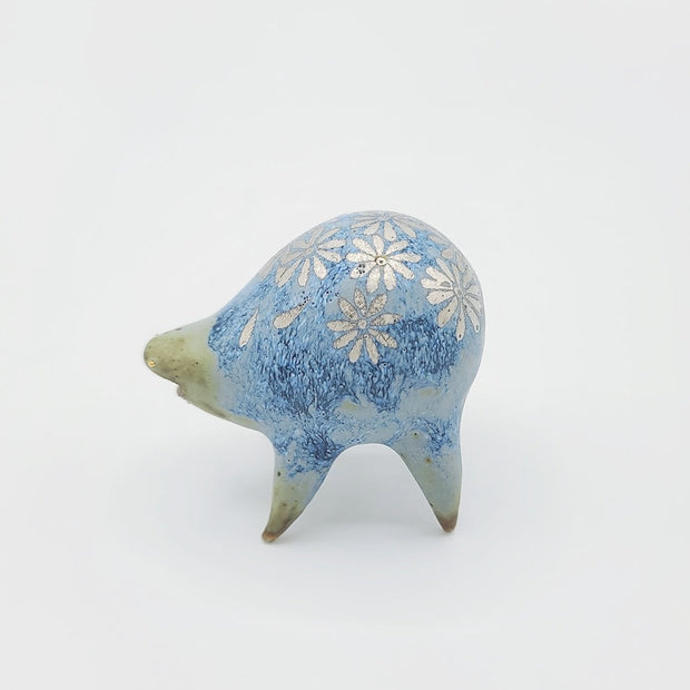 Rounded sage blue ceramic sculpture of a quadruped with short legs and small silver eyes over a closed mouth. Its body features a silver daisy pattern.