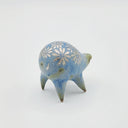 Rounded sage blue ceramic sculpture of a quadruped with short legs and small silver eyes over a closed mouth. Its body features a silver daisy pattern.