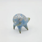 Rounded sage blue ceramic sculpture of a quadruped with short legs and small silver eyes over a closed mouth. Its body features a silver daisy pattern.