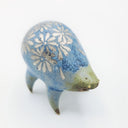 Rounded sage blue ceramic sculpture of a quadruped with short legs and small silver eyes over a closed mouth. Its body features a silver daisy pattern.