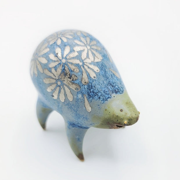 Rounded sage blue ceramic sculpture of a quadruped with short legs and small silver eyes over a closed mouth. Its body features a silver daisy pattern.