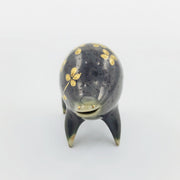 Rounded dark greenish blue ceramic sculpture of a quadruped with short legs and small gold eyes over a goofy open mouth smile. Its body features a gold flower pattern.