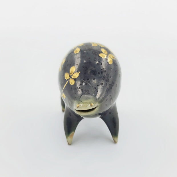 Rounded dark greenish blue ceramic sculpture of a quadruped with short legs and small gold eyes over a goofy open mouth smile. Its body features a gold flower pattern.