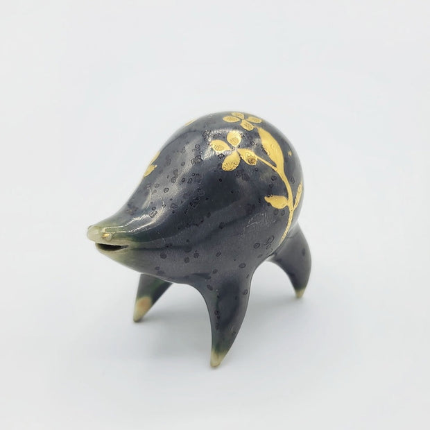 Rounded dark greenish blue ceramic sculpture of a quadruped with short legs and small gold eyes over a goofy open mouth smile. Its body features a gold flower pattern.