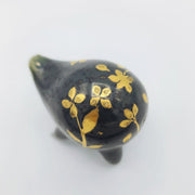 Rounded dark greenish blue ceramic sculpture of a quadruped with short legs and small gold eyes over a goofy open mouth smile. Its body features a gold flower pattern.