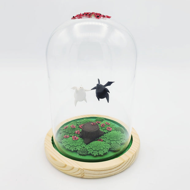 Series of delicate sculptures contained within a glass cloche. Atop the glass dome is a series of small red flowers. Floating down from the top are 2 chubby figures with wings, one white with angel wings and the other black with bat wings. They float over a green base with a tree stump and various foliage and more red flowers around it.