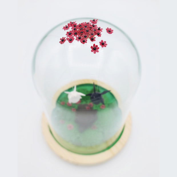 Series of delicate sculptures contained within a glass cloche. Atop the glass dome is a series of small red flowers. Floating down from the top are 2 chubby figures with wings, one white with angel wings and the other black with bat wings. They float over a green base with a tree stump and various foliage and more red flowers around it.