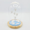 Series of delicate sculptures contained within a glass cloche. Atop the glass dome is a series of white clouds and floating around within are small white chubby creatures with angel wings. At the bottom is a blue base with white fluffy clouds sculpted.
