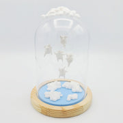 Series of delicate sculptures contained within a glass cloche. Atop the glass dome is a series of white clouds and floating around within are small white chubby creatures with angel wings. At the bottom is a blue base with white fluffy clouds sculpted.