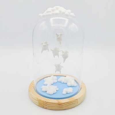 Series of delicate sculptures contained within a glass cloche. Atop the glass dome is a series of white clouds and floating around within are small white chubby creatures with angel wings. At the bottom is a blue base with white fluffy clouds sculpted.