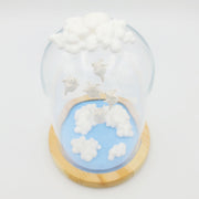 Series of delicate sculptures contained within a glass cloche. Atop the glass dome is a series of white clouds and floating around within are small white chubby creatures with angel wings. At the bottom is a blue base with white fluffy clouds sculpted.