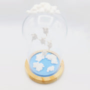 Series of delicate sculptures contained within a glass cloche. Atop the glass dome is a series of white clouds and floating around within are small white chubby creatures with angel wings. At the bottom is a blue base with white fluffy clouds sculpted.