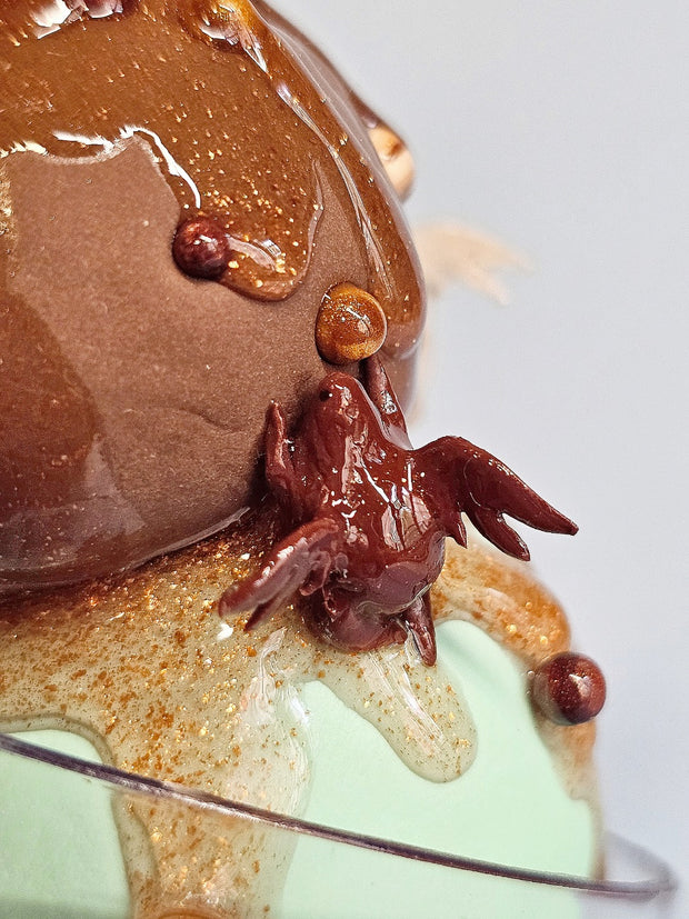 Sculpture of a triple scoop sundae in a cup of chocolate, mint and swirl. Sparkly sauce drips down it and nuts and small winged creatures adorn it.