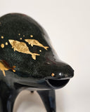Black ceramic sculpture of a rounded body quadruped creature with an open mouth goofy smile. It has golden fish silhouettes swimming on its back, with gold splatters as well.