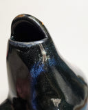 Black ceramic sculpture of a rounded body quadruped creature with an open mouth goofy smile. It has golden fish silhouettes swimming on its back, with gold splatters as well.