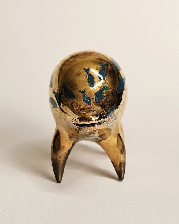 Gold ceramic sculpture of a rounded body quadruped creature with an open mouth goofy smile. It has blue fish silhouettes swimming on its back.