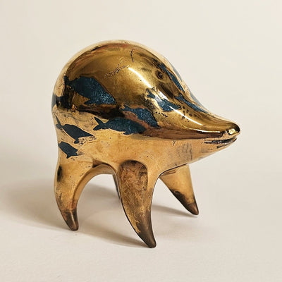 Gold ceramic sculpture of a rounded body quadruped creature with an open mouth goofy smile. It has blue fish silhouettes swimming on its back.