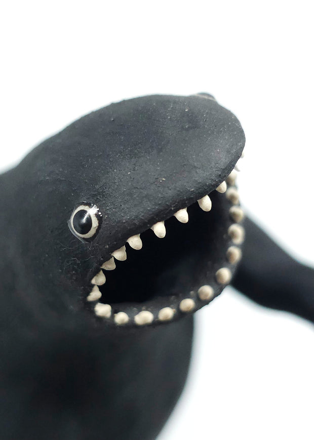 Black matte ceramic sculpture of a large Godzilla character, with many blue glazed spikes atop its back and a thick tail, standing with its mouth open. Its large tail rests on top of a small sitting dog. 
