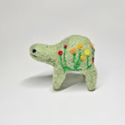 Needle felted wool sculpture of a light green rounded body quadruped creature with a cute smile and small black eyes. It has red, orange and yellow poppy flowers on its body.