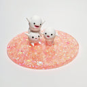 Sculpture of 3 small white characters with round bodies, tiny arms and legs, and cute cartoon expressions. They stand atop a pink glittery circular base.
