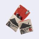 Rectangular textile with a pocket, the top can be folded over to make it into a pouch. Interior is bright orange and exterior is beige with designs of a triangular tiled roof with a clothesline and a red demon hanging off of it. Shown folded and unfolded.