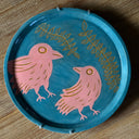 Blue ceramic plate with an inlayed illustration of a pink bird looking up and off to the side towards a 2nd, larger pink bird which looks back at it. Atop are 2 long vines with many leaves.