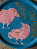 Blue ceramic plate with an inlayed illustration of a pink bird looking up and off to the side towards a 2nd, larger pink bird which looks back at it. Atop are 2 long vines with many leaves.