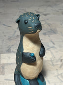 Ceramic sculpture of a dark grayish blue otter, standing on a small blue mound. It has a cream colored underbelly, black eyes, a cute face and holds a small teal shiny rock.