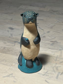 Ceramic sculpture of a dark grayish blue otter, standing on a small blue mound. It has a cream colored underbelly, black eyes, a cute face and holds a small teal shiny rock.