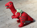 Ceramic sculpture of a red dinosaur wearing a green backpack that says 'GR Japantown"