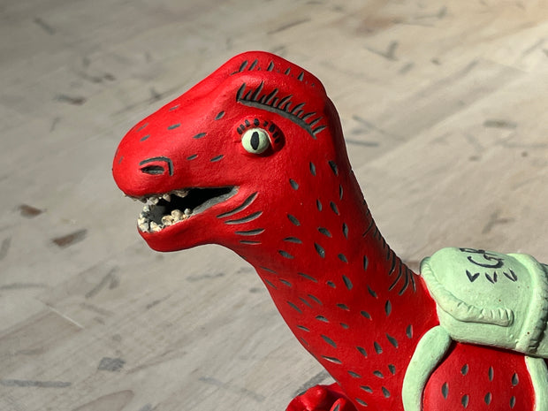 Ceramic sculpture of a red dinosaur wearing a green backpack that says 'GR Japantown"