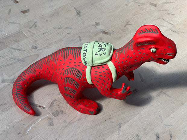 Ceramic sculpture of a red dinosaur wearing a green backpack that says 'GR Japantown"