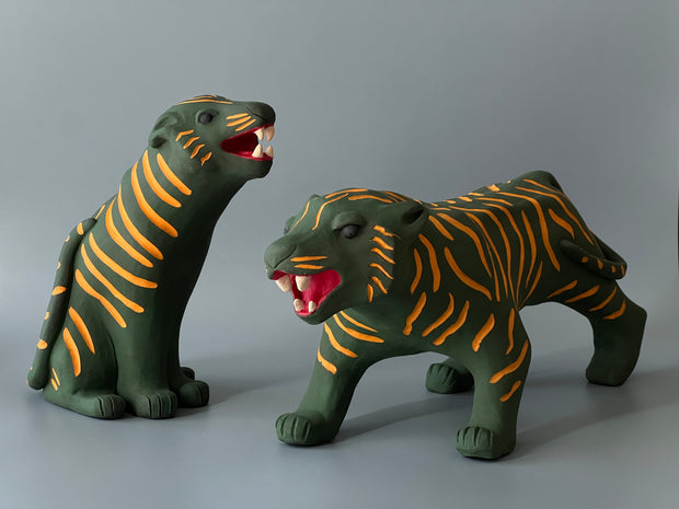 Matte finish ceramic sculpture of a green tiger with orange stripes, sitting on its hindlegs and with an open mouth smile. It sits next to a matching tiger, in a prowling stance.