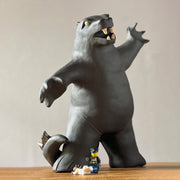 Ceramic sculpture of a large dark gray Godzilla style monster, with his mouth ajar and arms outspread. At his feet stand tiny lego characters.