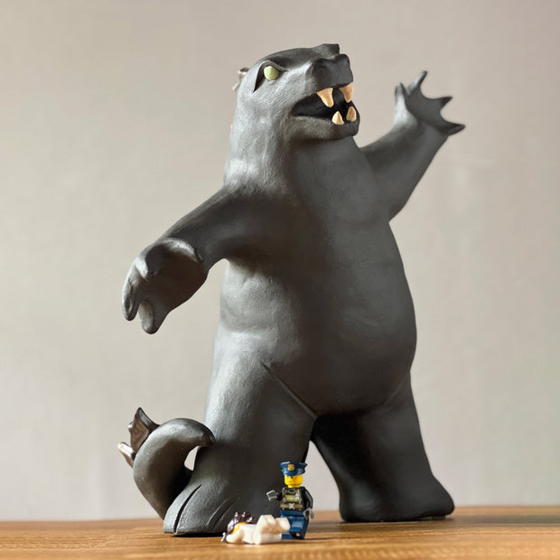 Ceramic sculpture of a large dark gray Godzilla style monster, with his mouth ajar and arms outspread. At his feet stand tiny lego characters.