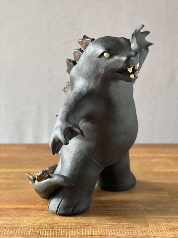 Ceramic sculpture of a large dark gray Godzilla style monster, with his mouth ajar and arms outspread. 