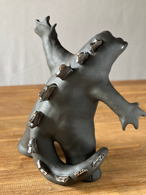 Ceramic sculpture of a large dark gray Godzilla style monster, with his mouth ajar and arms outspread. At his feet stand tiny lego characters.