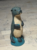 Ceramic sculpture of a dark grayish blue otter, standing on a small blue mound. It has a cream colored underbelly, black eyes, a cute face and holds a small teal shiny rock.